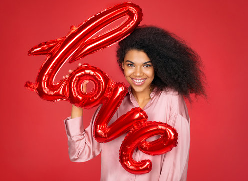 Smiling girl holds letter shaped balloon. Photo of young girl in love on red background. Valentine's Day
