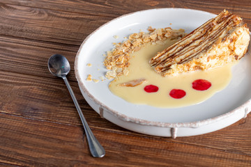 Dessert Napolion with custard, condensed milk sauce, crumbs of dough and raspberry jam. In the background is a cappuccino. On a wooden natural background.
