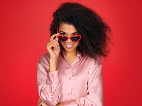 Cute girl in stylish glasses in the shape of hearts looks into camera. Photo of attractive latin girl in love on red background