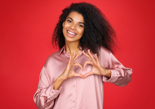 Beautiful girl shows heart symbol, shapes love sign with hands. Photo of african american girl in love on red background. Be my Valentine