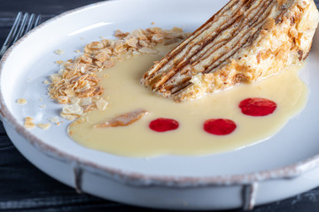 Napoleon dessert with custard, condensed milk sauce, crumbs of dough and raspberry jam. In the background is a cappuccino. On a wooden natural background.