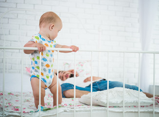 Cute little baby is standing on the edge of the bed holding on to the headboard against the background of his sleeping parents. Concept of tired about happy newly made parents. Copyspace