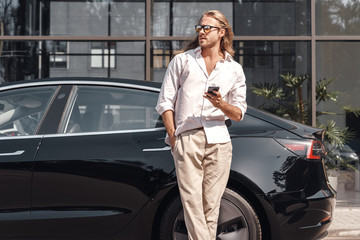 Mode of Transport. Man with long hair traveling by electric car leaning back browsing smartphone...