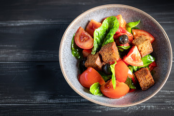 Italian salad of sliced tomatoes, basil leaves, olives, large pieces of croutons. In a deep plate, on a dark background.