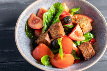 Italian salad of sliced tomatoes, basil leaves, olives, large pieces of croutons. In a deep plate, on a dark background.