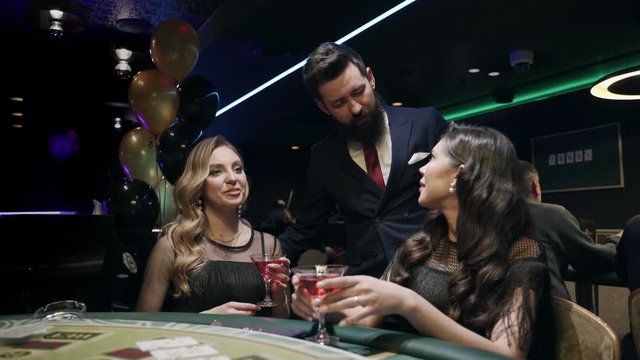Casino: A bearded man communicates with preatty girls and they smile and laugh. Аlcoholic red champagne in the hands. They are sitting at the pocker table