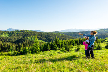 Fototapeta na wymiar Young mother and two little daughters travelers stand on a slope with a gorgeous view of the hills covered with dense fir forest against the blue sky on sunny warm summer day. Family tourism concept