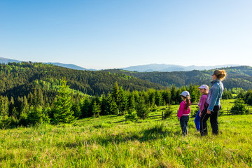 Young mother and two little daughters travelers stand on a slope with a gorgeous view of the hills covered with dense fir forest against the blue sky on sunny warm summer day. Family tourism concept