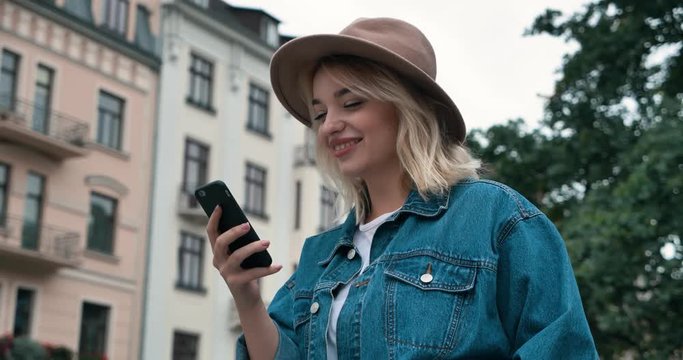 Portrait of Beautiful Blonde woman walking in the Town while Typing on Smartphone’s screen. Looking stylish wearing Jeans Jacket and Hat. Having hand in the Pocket. Girl with Charming smile.