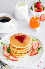 A stack of pancakes with a heart of jam on top with fresh strawberries and mint on a white plate on a light background. Breakfast for Valentine's Day. Vertical orientation. Copy space