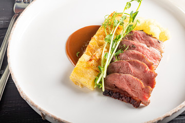Duck breast with a side dish of potato casseroles, seasoned with pea microgreen with tomato and chili sauce. On a white plate. On a wooden background,