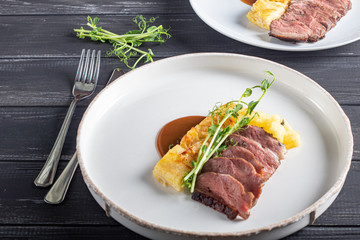 Duck breast with a side dish of potato casseroles, seasoned with pea microgreen with tomato and chili sauce. On a white plate. On a wooden background,
