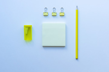 Top view of stationery in yellow and blue colours: three clips, sharpener, pencil and sticky notes on white background. Concept of education, business, minimalism.