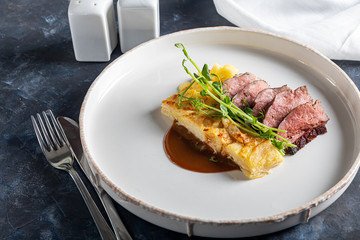 Duck breast with a side dish of potato casseroles, seasoned with pea microgreen with tomato and chili sauce. On a white plate. On a wooden background,