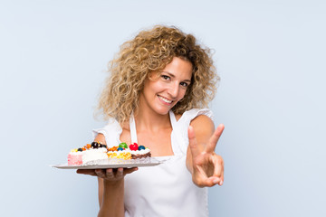 Young blonde woman with curly hair holding lots of different mini cakes smiling and showing victory sign