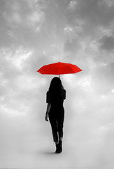 Silhouette of a girl with red umbrella walking under a cloudy sky. Surreal atmosphere and contrast with the full red color of the umbrella and the gray background