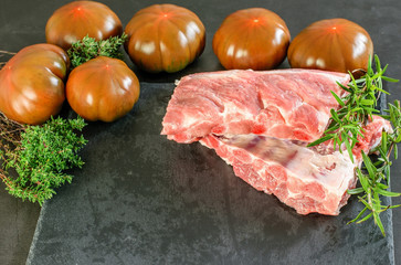 Pieces of uncooked fresh pork ribs with tomatoes and herbs on the black kitchen background, prepared for cooking.
