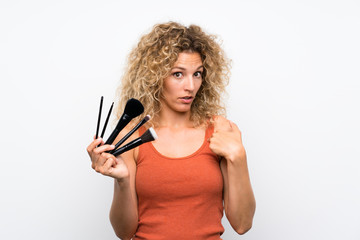 Young blonde woman with curly hair holding a lot of makeup brush with surprise facial expression