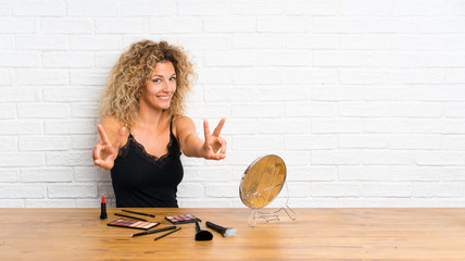 Young woman with lots of makeup brush in a table smiling and showing victory sign