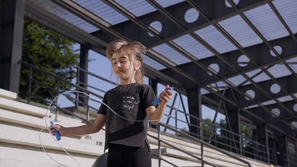Attractive portrait of smiling sporty little girl in black sport clothes which jumping with skipping rope on the platform of the stadium - Powered by Adobe