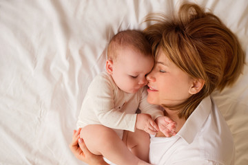 mother plays with a baby of 6 months of European appearance, both in white clothes are lying on the bed