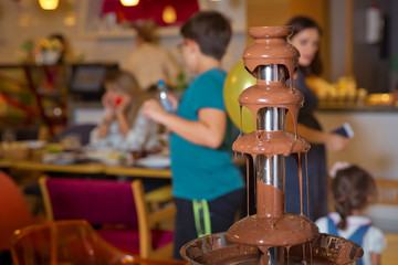 Obraz premium Chocolate fountain with fruits. Children birthday party . Homemade chocolate fountain fondue with marshmallow on a skewer dripping in chocolate sauce on blurred background and copy space .