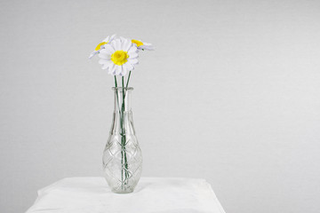 drei weiß gelbe gebastelte Papierblumen mit grünem stil in Glasvase mit Ornament vor weißem Tisch Platz für Text