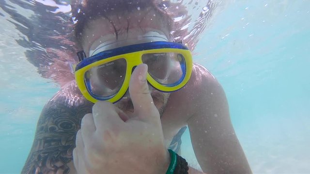 A young man swims underwater in the sea, On it are yellow-blue glasses for scuba diving, The sun's rays make their way through the water, He shows everything well with his finger.