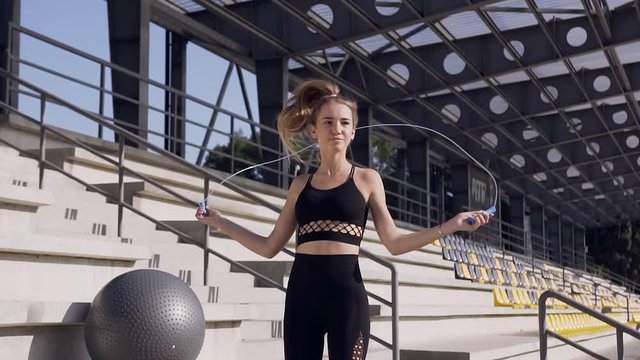 Front view of lovely slender sporty young woman in fitness clothes which doing jumping exercises with skipping rope on the tribune on the stadium