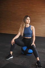 Fototapeta na wymiar Vertical full-length shot beautiful female athlete, sportswoman in activewear with long blond hair, sit on medicine ball, turn right and smiling, rest during workout break, keep body strong and fit