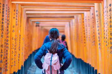 Young woman traveling at Fushimi Inari taisha Shrine, happy asian traveler looking vibrant orange torii gates. landmark and popular for tourists attractions in Kyoto. Japan, 27 November 2019