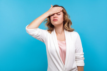 Facepalm. Portrait of forgetful woman with wavy hair in white jacket slapping hand on forehead, feeling sorrow regret about forgotten event, bad memory. indoor studio shot isolated on blue background