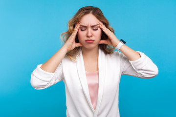 Fototapeta premium Headache. Portrait of unhealthy girl with wavy hair in white jacket suffering migraine, standing with closed eyes and touching head to relieve pain, feeling sick stressed. studio shot, blue background