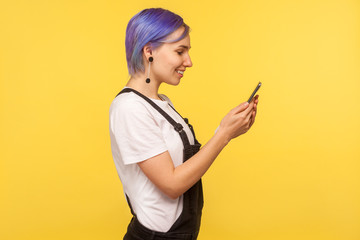 Side view of glad hipster girl with violet hair in overalls using cell phone, texting or reading message with good news, smiling enjoying mobile app. isolated on yellow background, indoor studio shot