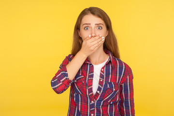 I won't tell anyone! Portrait of scared ginger girl in checkered shirt covering mouth with hand and looking frightened at camera, intimidated female afraid to talk. studio shot, yellow background