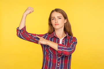Look, I'm strong woman! Portrait of self-confident ginger girl in casual shirt pointing at biceps,...