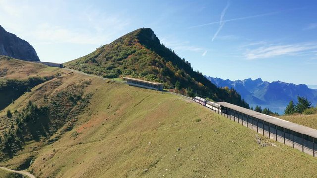 Follow aerial shot of unique switzerland railway train going uphill through tunels in mountains. Green grass with blue sky. Cableway lift for skiers at background