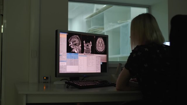 In dark room in hospital laboratory technician looks at results of studies, on monitor are pictures of patient’s brain, tomographic examination, disease search, diagnosis, preparation of results