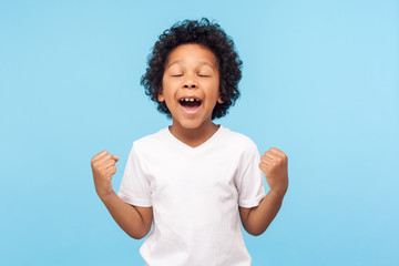 Child winning, success. Portrait of excited little boy in T-shirt raising hands and screaming delighted with victory and luck, expressing extreme joy. indoor studio shot isolated on blue background