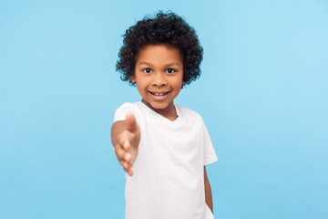 Portrait of friendly cheerful little boy with curly hair in white T-shirt giving hand to handshake and smiling at camera, looking gullible trustful. indoor studio shot isolated on blue background
