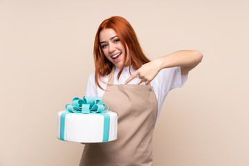 Redhead teenager girl with a big cake over isolated background and pointing it