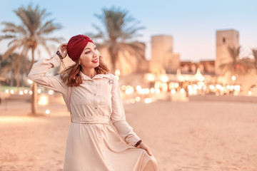 Happy asian woman in turban travels in Sahara desert. Oasis and Bedouin village at the background. Adventure and life experience concept