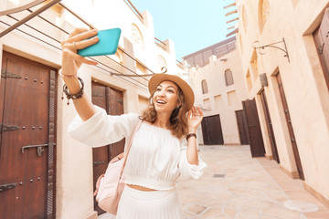 Happy asian girl in white dress taking selfie against narrow streets of old Arabian town in Dubai. Travel destinations and tourism in UAE concept