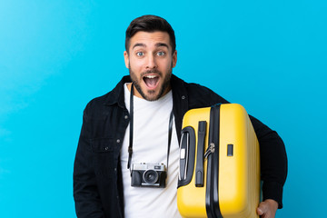 Traveler man holding a suitcase over isolated blue background with surprise and shocked facial expression