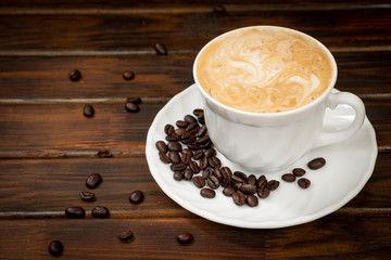 Cappuccino in a white glass on a saucer. Coffee beans on a saucer. Brown background