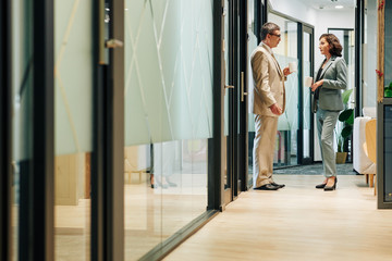 Middle-aged man and mature woman chatting during coffee break standing in office building corridor, horizontal long shot