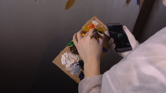 Artist designer draws an eagle on wall. Craftsman decorator paints picture with acrylic oil color looking at sketch in phone. Painter painter dressed in paint coat. Indoor. Dark magic cinematic look.