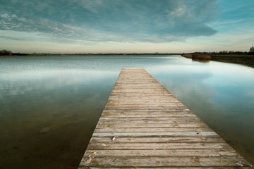 Fototapeta na wymiar Long wooden bridge on the calm lake, evening clouds on the sky