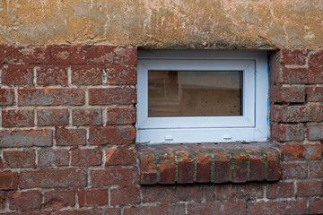 Modern white plastic window with a glare leading to the basement with a brick shelf under it in the lower part, lined with brown broken off in places bricks, on the beige facade of an old shabby house