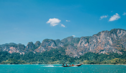 Wooden motorboat vessel using a long propeller shaft sailing with passengers on Thai Khao Sok Lake with foresty mountains around. Exotic countries traveling concept image.